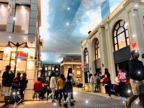 A bustling indoor street scene with shops, people walking, and a painted sky ceiling.