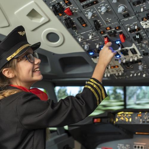 A young girl in a pilot's uniform smiles while interacting with the controls in a flight simulator cockpit.