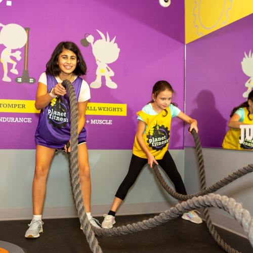 Two girls are exercising with battle ropes in a colorful gym, smiling and engaged in their workout.