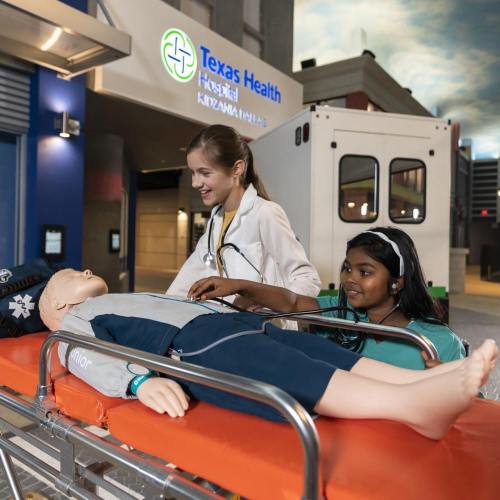 A medical professional and a child assist a mannequin on a stretcher at Texas Health hospital.
