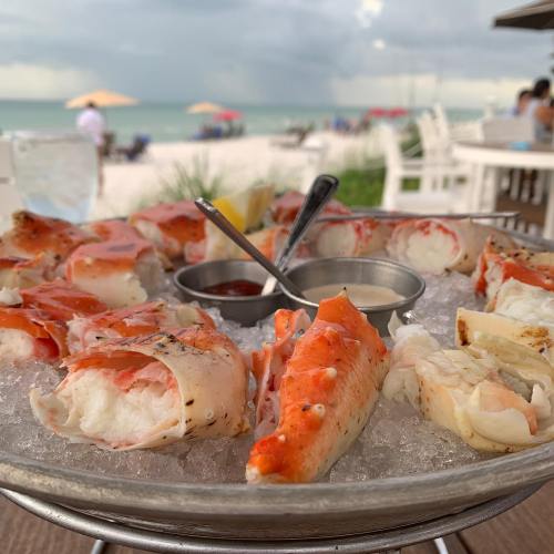 A seafood platter with crab legs on ice, served with dipping sauces, against a beach backdrop.