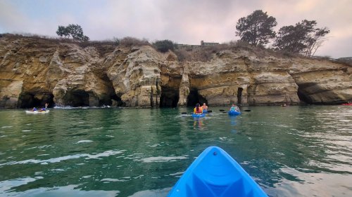 Kayakers explore a rocky coastline with caves, surrounded by calm water and a cloudy sky.