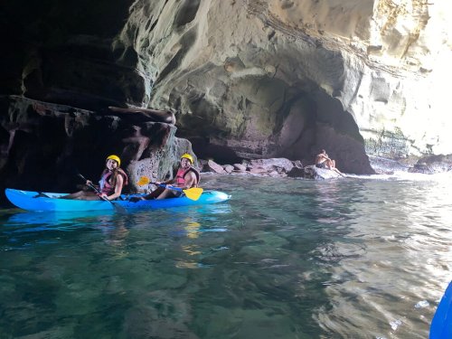 Two people in kayaks explore a rocky cave, while another person relaxes on a rock nearby.