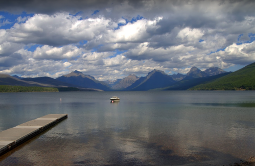 A serene lake surrounded by mountains under a cloudy sky, with a small boat floating near a wooden dock.