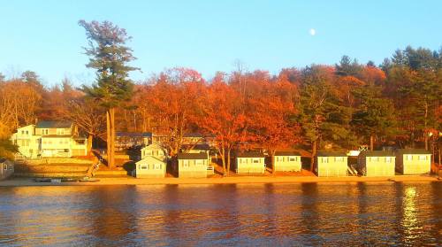 Cottages along a lake shore, surrounded by autumn trees and a clear blue sky with a hint of moon.