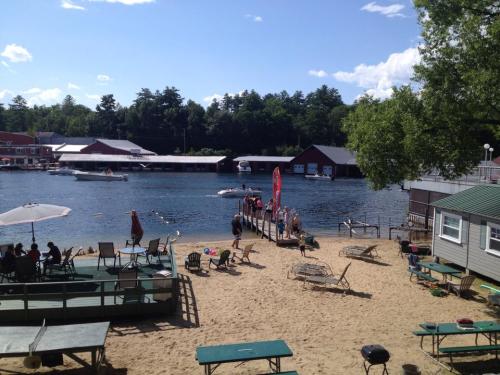 A sunny lakeside scene with sandy beach, dock, boats, and people enjoying the water and relaxing on chairs.