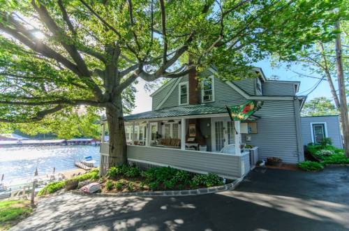 A charming gray house with a porch, surrounded by trees, near a lake on a sunny day.