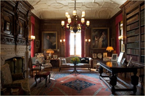 A cozy, elegant library with wooden walls, vintage furniture, paintings, and a chandelier, illuminated by warm light.