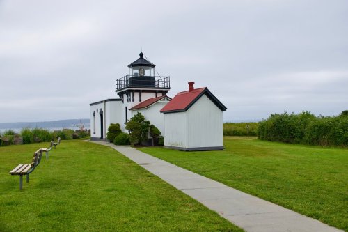 A lighthouse with a black and white tower, surrounded by green grass and benches, under a cloudy sky.