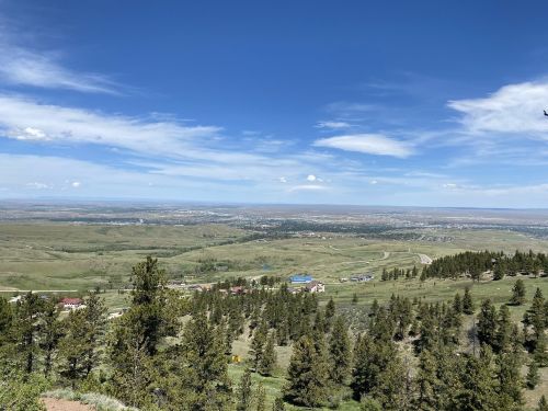 A panoramic view of rolling green hills, trees, and a distant town under a clear blue sky with scattered clouds.