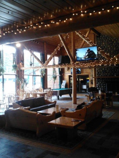 Cozy lodge interior with wooden beams, soft seating, and warm lighting, featuring a fireplace and large windows.