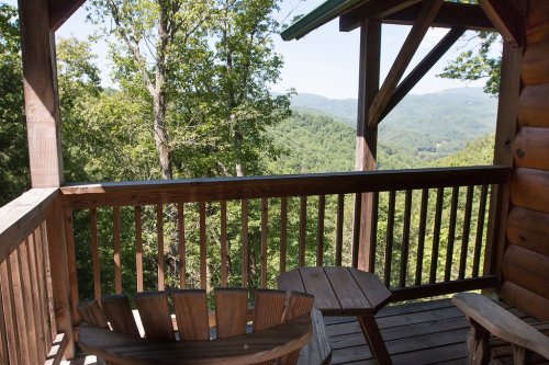 A wooden balcony with two chairs overlooks a lush green mountain landscape under a clear blue sky.