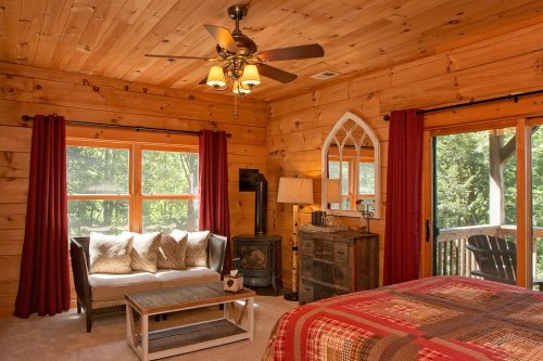Cozy cabin bedroom with wooden walls, a sofa, large windows, and a plaid bedspread, featuring warm lighting and rustic decor.