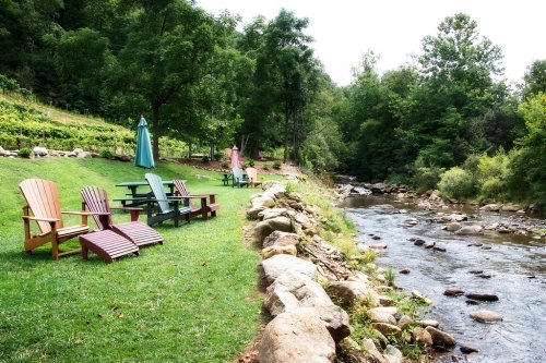 Colorful Adirondack chairs line a grassy bank beside a gently flowing stream, surrounded by lush trees.