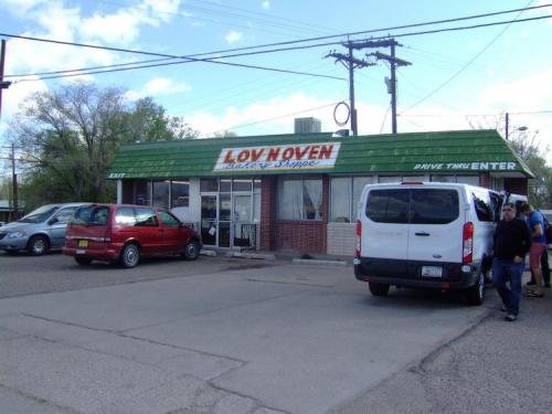 A small bakery shop with a green roof, parked cars, and people outside on a sunny day.