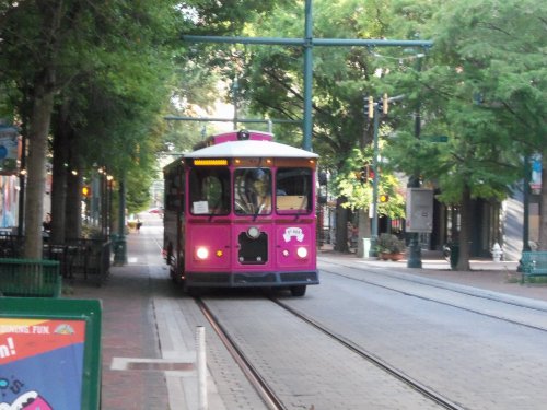 A pink trolley car travels down a tree-lined street, surrounded by greenery and urban scenery.