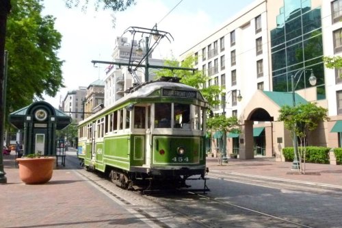 A green vintage tram on a city street, surrounded by trees and modern buildings.