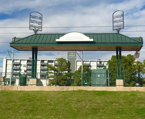 A green train station shelter with musical notes on top, surrounded by grass and buildings in the background.