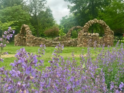 A stone ruin with archways surrounded by lush greenery and blooming purple flowers in the foreground.