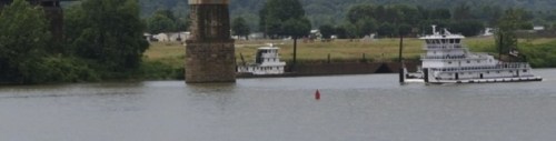 Two boats navigate a river under a bridge, with green hills and fields in the background.