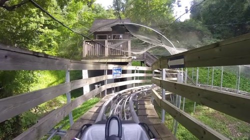 A view from a roller coaster track winding through trees, approaching a wooden structure in the background.