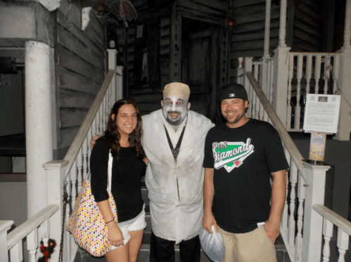 A man in a creepy costume poses with two smiling people in front of a haunted house entrance.
