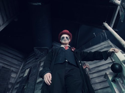 A person in a top hat and coat stands ominously in a dimly lit, eerie setting with a haunted house backdrop.