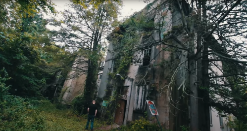 A person stands in front of an abandoned, overgrown building surrounded by trees and foliage.
