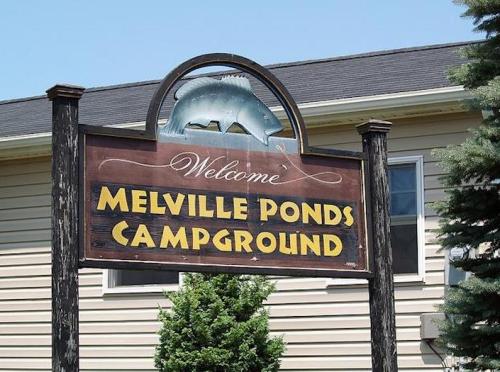 Sign for Melville Ponds Campground, featuring a fish graphic and welcoming text, set against a building backdrop.