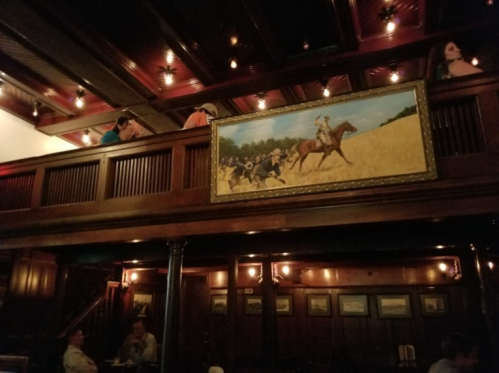 A dimly lit restaurant interior featuring wooden decor, a large painting of horse riders, and patrons seated at tables.