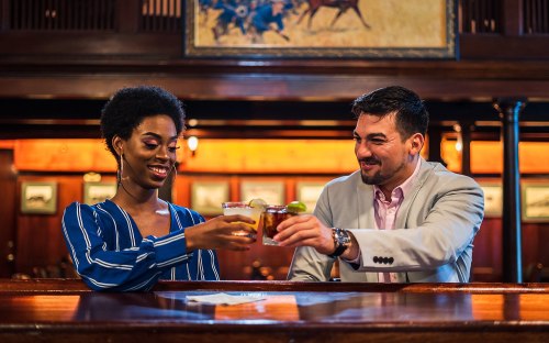 A man and woman clink glasses at a bar, smiling and enjoying drinks together in a warm, inviting atmosphere.