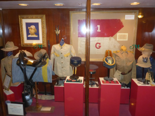 Display of historical military uniforms, hats, and memorabilia in a glass case, featuring various artifacts and decorations.