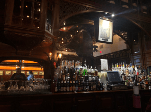 A dimly lit bar with an ornate wooden design, showcasing a variety of liquor bottles and glassware.