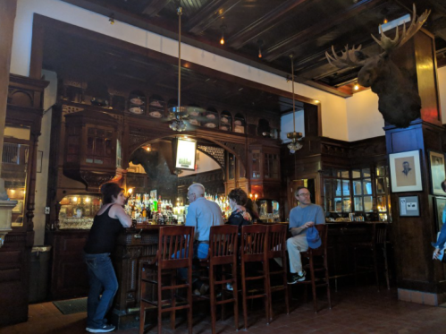 A cozy bar scene with patrons sitting at the counter, surrounded by wooden decor and a mounted moose head.