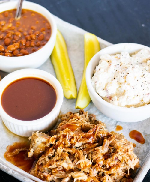 A tray with pulled pork, barbecue sauce, pickles, baked beans, and potato salad.