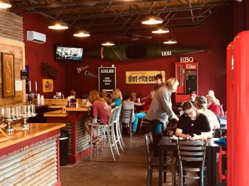 A lively restaurant interior with patrons dining, a bar area, and vintage decor, including a TV and a red Coca-Cola fridge.