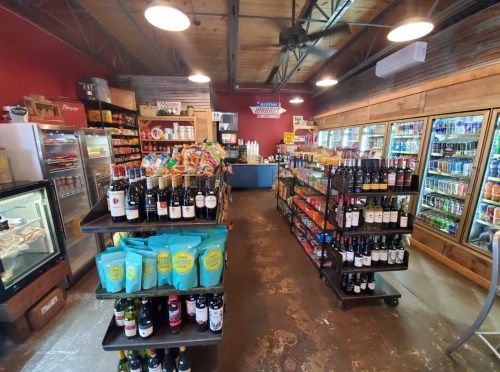 A well-stocked convenience store with shelves of snacks, drinks, and wine, featuring a rustic interior and bright lighting.