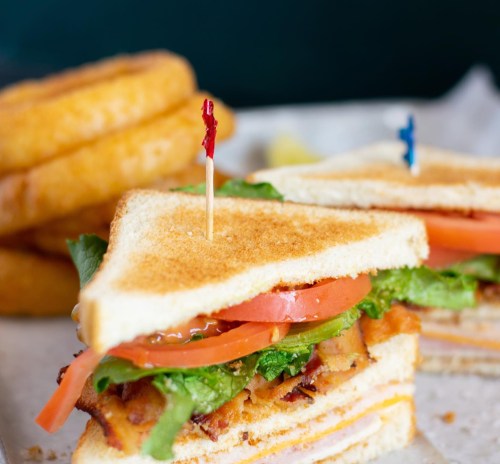 A close-up of a toasted sandwich with lettuce, tomato, and bacon, served with onion rings in the background.