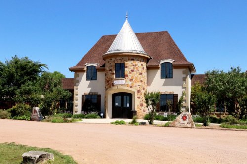 A charming stone and stucco building with a conical roof, surrounded by greenery and a gravel pathway.
