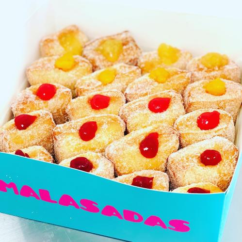 A box of malasadas, golden-brown doughnuts topped with colorful fruit jelly, arranged neatly.