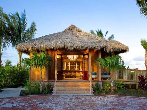 A tropical hut with a thatched roof, surrounded by palm trees, featuring a wooden deck and inviting entrance.