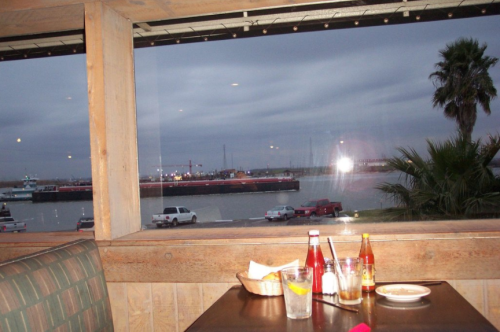 A restaurant table with drinks and condiments, overlooking a cloudy waterfront view with a barge in the distance.