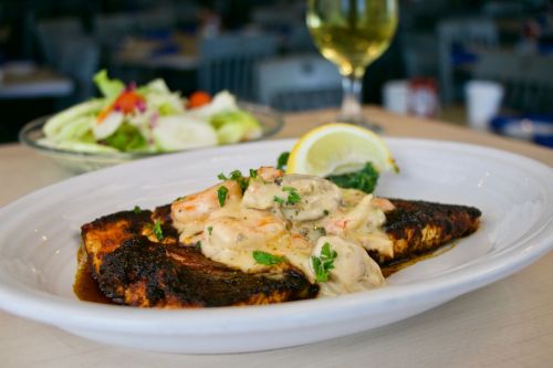 A plate of blackened fish topped with shrimp and creamy sauce, served with a lemon wedge and a side salad.