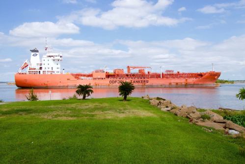 An orange tanker ship docked near a grassy shore under a blue sky with scattered clouds.