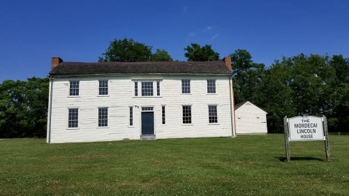 Historic white house with a sign reading "The Mordecai Lincoln House," set against a clear blue sky and green grass.