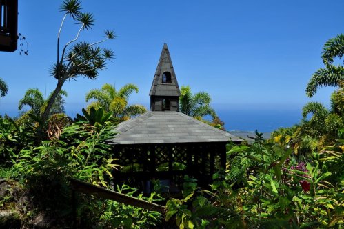 A small, peaked-roof structure surrounded by lush greenery and palm trees, overlooking a blue ocean under a clear sky.
