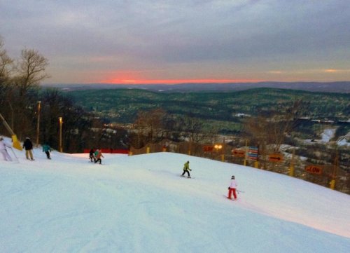 Skiers descend a snowy slope at sunset, with a scenic view of hills and trees in the background.