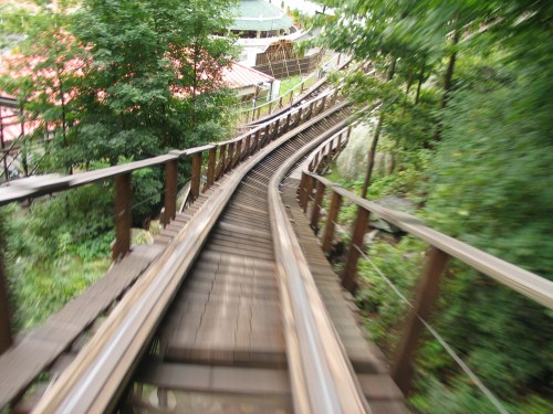 A winding wooden roller coaster track surrounded by lush greenery, creating a sense of speed and excitement.