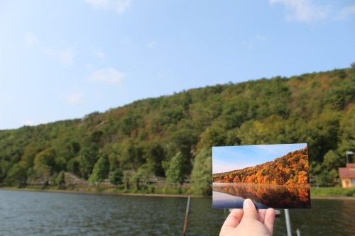 A hand holds a postcard of a colorful autumn landscape over a serene lake, with a green hillside in the background.