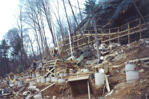 Construction site with workers building a wooden structure on a hillside, surrounded by trees and rocky terrain.